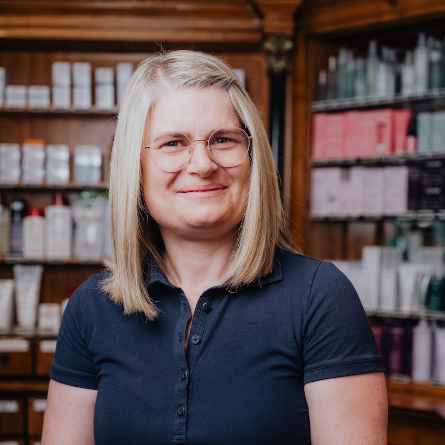 Eine lächelnde Person mit Brille und blondem Haar steht vor einem Regal mit Kosmetikprodukten in einem Geschäft.