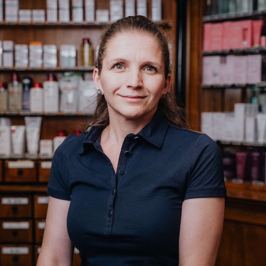 Eine Frau mit dunkelblauem Hemd steht vor einem Regal mit Kosmetikprodukten und schaut lächelnd in die Kamera.