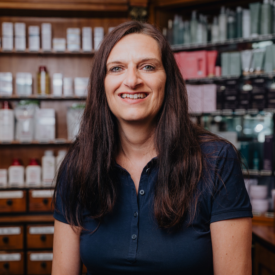 Eine lächelnde Frau mit langen braunen Haaren trägt ein dunkelblaues Polohemd und steht vor einem Regal mit Kosmetikprodukten.