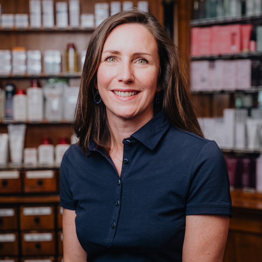 Eine lächelnde Frau mit braunen Haaren trägt ein dunkelblaues Poloshirt und steht vor einem Regal mit verschiedenen Kosmetikprodukten.
