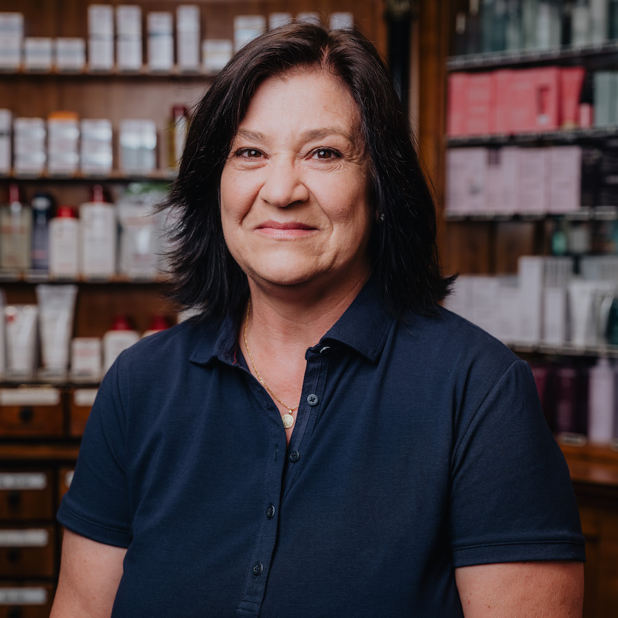 Eine Frau mittleren Alters mit dunklem Haar und dunkelblauem Polohemd steht vor einem Regal mit Kosmetikprodukten.