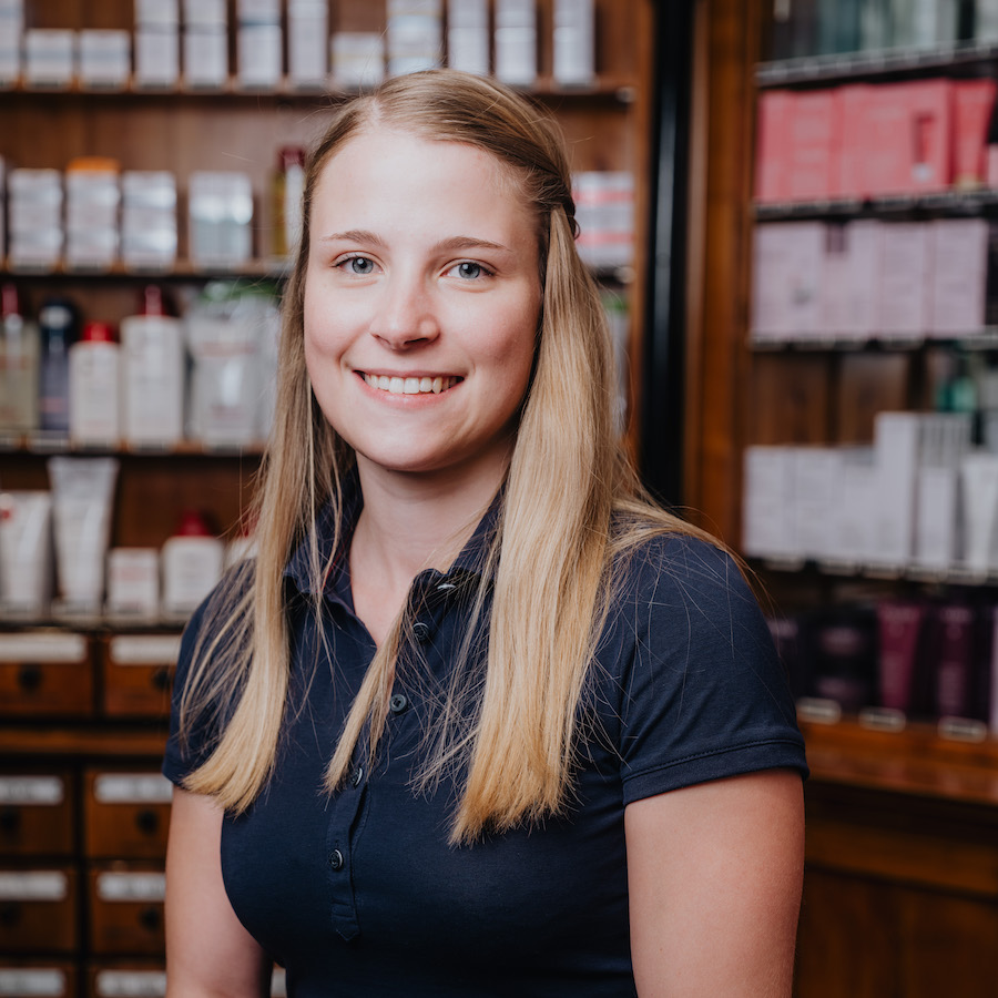 Eine lächelnde Frau mit langen blonden Haaren trägt ein dunkelblaues Poloshirt und steht vor einem Regal mit Pflegeprodukten.
