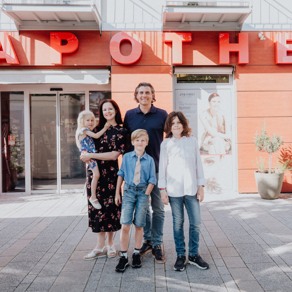 Eine Familie steht lächelnd vor einer Apotheke. Drei Kinder, zwei Jungs und ein Mädchen, mit ihren Eltern in sommerlicher Kleidung.