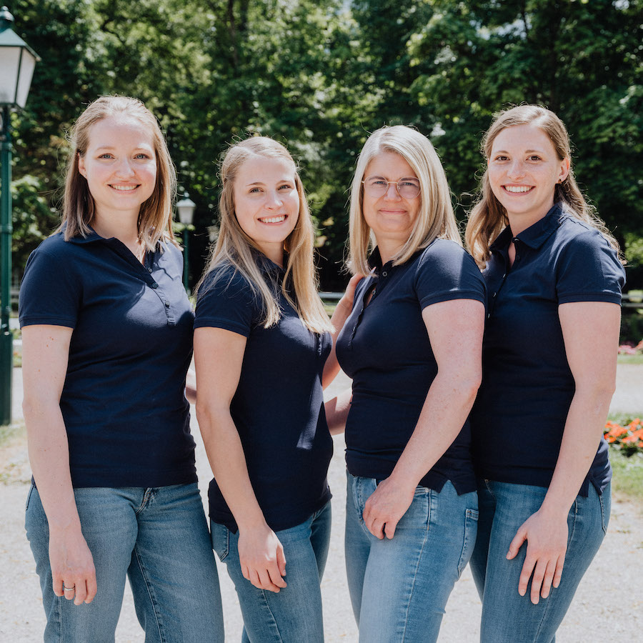 Vier mitarbeitende Frauen der Stadt-Apotheke Bad Hall. Alle tragen dunkelblaue Poloshirts und Jeans, im Hintergrund sind Bäume zu sehen.
